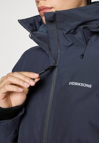 Persona, kas velk augšā rāvējslēdzēju tumši zilā Didriksons jaciņā ar augstu apkakli un auklu detaļām apkakles tuvumā.