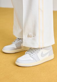 Bílé tenisky Nike s šedými akcenty, texturovanými panely a krémovou gumovou podrážkou; spárované s bílými kalhotami s bočními pruhy a detailem loga.