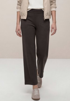 Femme portant un pantalon noir ample, une veste beige, un pull blanc et des baskets beige marchant vers l'avant sur un fond clair uni.