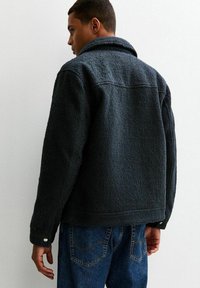 Czarna polarowa kurtka o teksturowanym wykończeniu, z ostrym kołnierzem i guzikami przy mankietach, noszona na niebieskich dżinsach.