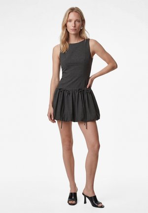 Kvinde iført ermeløs mørkegrå minikjole med samlet nederdel og sorte, åbne hæl-sandaler med hæl, stående med den ene hånd på hoften.