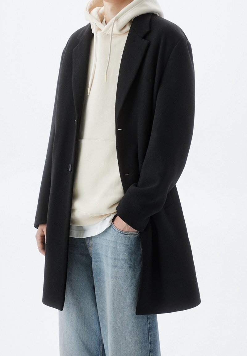 Cappotto lungo nero con design strutturato, indossato sopra un hoodie beige. Jeans in denim chiaro con una vestibilità rilassata, che mostrano un look a strati casual.