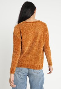 Pomarańczowy sweter o strukturalnej fakturze z długimi rękawami, prążkowanym dekoltem i rozciętymi brzegami. W połączeniu z jasnoniebieskimi dżinsami.