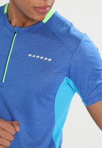Camisa deportiva de manga corta en azul con detalles en azul claro. Cuenta con un medio cierre y el logo "DARE2B" en blanco en el pecho. Material elástico y transpirable.