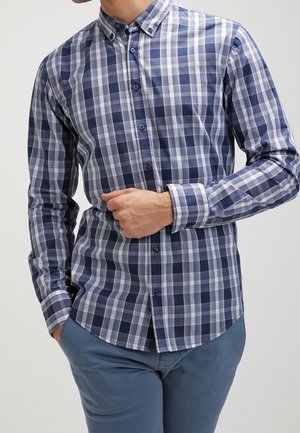 Mies, joka pitää päällään sinivalkoruudullista pitkähihnaista nappipaitaa ja siniset housut, käsi säätämässä paidan etuosaa.