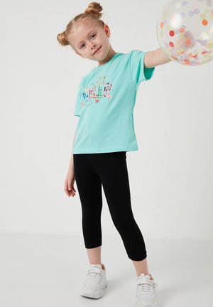 Jong meisje met blond haar in knotjes, gekleed in een turquoise shirt en zwarte legging, houdt een doorzichtige ballon met confetti vast tegen een witte achtergrond.