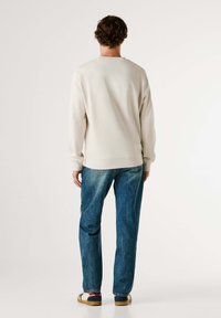 Felpa beige con polsini e orlo a coste, abbinata a jeans blu sbiaditi. Modello con indosso scarpe casual dal design multicolore.