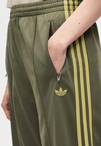 Χέρι στην τσέπη πράσινου λαδί παντελονιού Adidas με κίτρινες ρίγες, φερμουάρ και το λογότυπο τριφύλλι Adidas στο πλάι.
