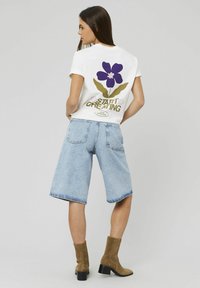 Femme aux longs cheveux noirs portant un t-shirt blanc avec une fleur violette et le texte "Commencez à créer", un short en jean clair et des bottines marron.