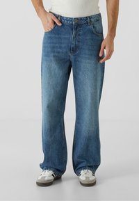 Modri denim kavbojki z ohlapnim krojem, ki imajo pet žepov in gumbasto zapiranje v pasu. Nosi se z belimi in sivoi športnimi copati.
