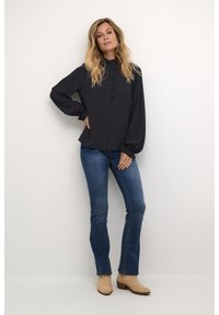 Black blouse with ruffle detailing along the button placket, long sleeves with gathered cuffs, paired with dark blue jeans and beige boots.