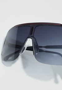 Occhiali sportivi da sole con montatura nera, lenti grigie e un accentuante superiore riflettente rosso. Presentano un design avvolgente con un ponte nasale distintivo.