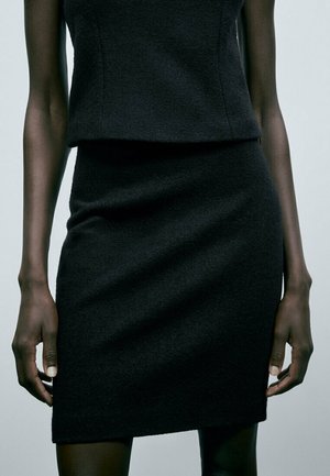 Femme portant un haut noir ajusté sans manches et une jupe noire assortie, debout les bras détendus le long du corps devant un fond uni.