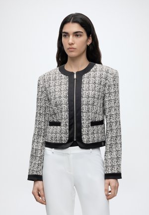 Vrouw met een zwart-witte tweedjas met rits aan de voorkant, zwarte bies en witte broek, staand tegen een egale achtergrond.