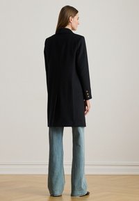 Μαύρο μάλλινο παλτό με ραμμένη σχεδίαση, που διαθέτει γιακά με εγκοπές και χρυσά κουμπιά στα μανίκια, συνδυασμένο με ανοιχτό μπλε μπατζάκια με φαρδιά άκρα.