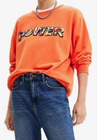 Oranje sweatshirt met meerkleurige geborduurde "POWER" tekst, gecombineerd met blauwe spijkerbroek. Zilveren ketting als accessoire.