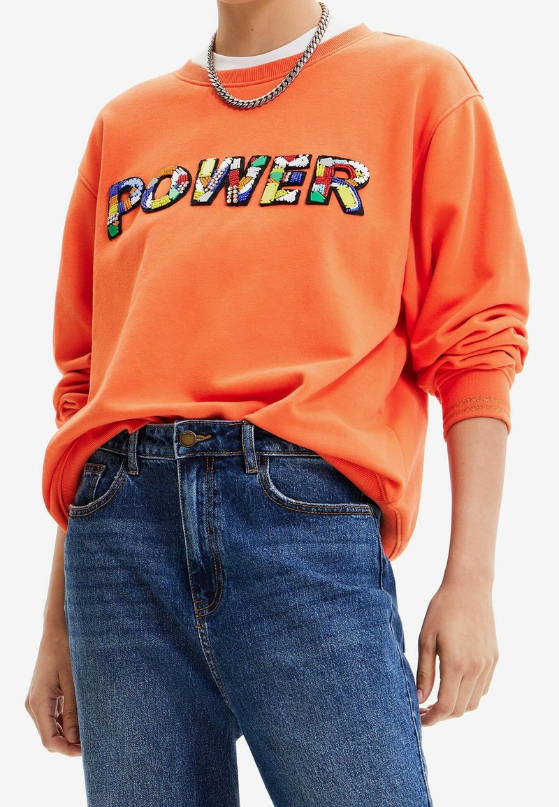 Oranje sweatshirt met meerkleurige geborduurde "POWER" tekst, gecombineerd met blauwe spijkerbroek. Zilveren ketting als accessoire.