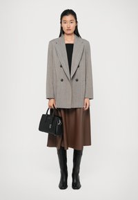 Blazer gris à double boutonnage par-dessus un haut noir, jupe marron taille haute, sac à main noir et bottes noires jusqu'aux genoux. Textures lisses et design structuré.