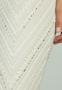 Robe blanche avec un motif en chevrons, présentant un tissu texturé et des perles décoratives éparpillées dans des tons dorés et argentés le long du motif.