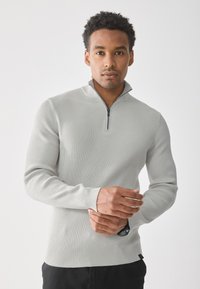 Hombre con suéter de canalé gris claro con cuello de media cremallera, de pie contra un fondo blanco, ajustándose la muñeca.