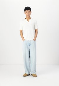 Polo de punto blanco de manga corta con escote en V, combinado con jeans de pierna ancha azul claro y zapatillas beige, de pie contra un fondo liso.