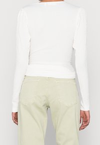Top bianco a maniche lunghe con texture e silhouette aderente, abbinato a pantaloni verdi chiari a vita alta con tasche posteriori.