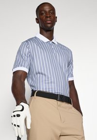 Polo com riscas em azul e branco, com colarinho branco e mangas enroladas. Calças beiges com um cinto preto. Usando uma luva de golfe branca.