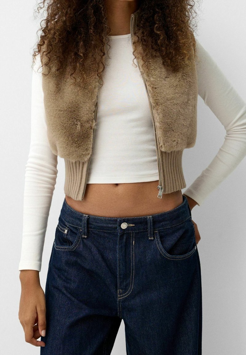 Beige Kunstfell-Cropped-Weste mit gerippten Kanten, Reißverschluss und dunklen Jeans. Langärmliges weißes Oberteil darunter.