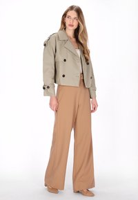 Khakifarbene kurze Jacke mit doppelt geknöpften schwarzen Knöpfen; kombiniert mit beigeweiten Hosen und passenden Wildleder-Absätzen.