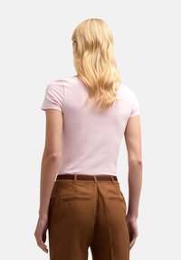 Jasnoróżowa dopasowana koszulka z krótkim rękawem, noszona z brązowymi spodniami z gładkim pasaem i dwiema tylnymi kieszeniami.