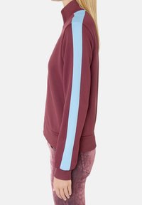 Vínová mikina s vysokým límcem, s námořnickými modrými postranními pruhy. Vyrobena z texturované tkaniny, s přiléhavým střihem a žebrovanými manžetami.