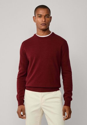 Uomo che indossa un maglione bordeaux a girocollo su una t-shirt bianca e pantaloni beige chiaro, in piedi contro uno sfondo grigio uniforme.