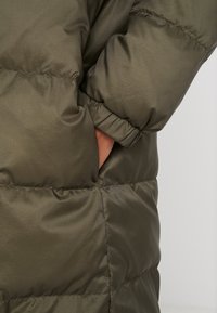 Olivgrön dunjacka med slät yta, utrustad med sidoficka och elastiska muddar för en bekväm passform. Quiltat mönster som tillför dimension.