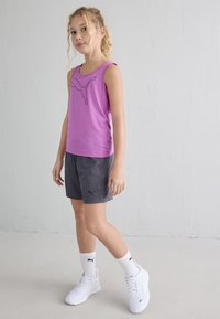 Noor tüdruk, kes kannab lillat Puma tank-tossu, halli lühikest püksikut, valgeid Puma sokke ja valgeid tossusid, seisab läbi lihtsa heleda seina.