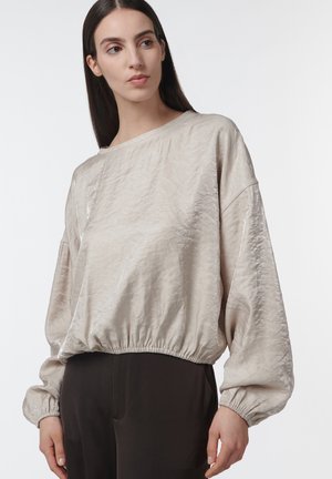 Mujer con cabello largo y oscuro, vestida con una blusa beige suelta y brillante con puños y dobladillo fruncidos, combinada con pantalones oscuros, sobre un fondo liso.
