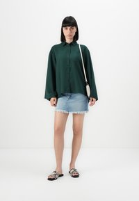 Kvinne iført en mørkegrønn oversized skjorte, lys blå slitt denimskjørt, sandaler i slangeskinnsutførelse og en liten hvit crossover-veske.