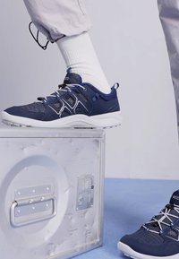 Scarpe sportive blu navy con tessuto in rete e sintetico, dotate di accenti bianchi, lacci e suola in gomma testurizzata, posizionate su una scatola argentata.