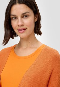 Oranger Cardigan mit strukturiertem Gewebe, übereinander getragen mit einem glatten orangenen T-Shirt. Der Rundhalsausschnitt ist rund, und der Cardigan hat ein offenes Frontdesign.