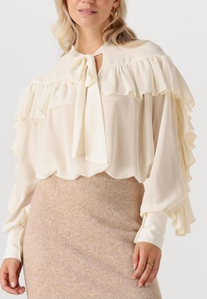 Woman wearing a cream ruffled blouse with a bow at the neck and long sleeves paired with a beige wool skirt.