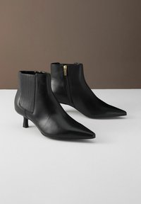 Černé kožené kotníkové boty s špičatou špičkou, s žebrovanými bočními panely a zlatým zipem jako akcentem. Blokový podpatek pro stabilitu.