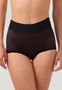 Vrouw in doorschijnende zwarte hooggesneden onderbroek, staande met ontspannen armen, weergegeven vanaf de taille tot halverwege de bovenbenen op een lichtgrijze achtergrond.