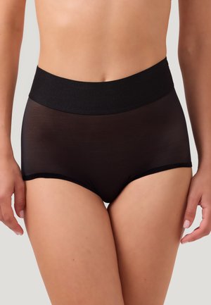 Vrouw in doorschijnende zwarte hooggesneden onderbroek, staande met ontspannen armen, weergegeven vanaf de taille tot halverwege de bovenbenen op een lichtgrijze achtergrond.