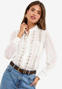 Vrouw met lang bruin haar, goudkleurige creolen, een witte kanten blouse met pofmouwen en een blauwe spijkerbroek met een bruine riem.