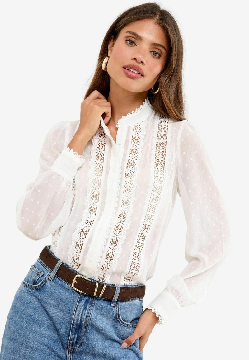 Vrouw met lang bruin haar, goudkleurige creolen, een witte kanten blouse met pofmouwen en een blauwe spijkerbroek met een bruine riem.