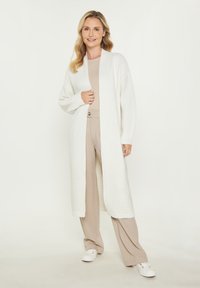 Cardigan lung alb din tricot texturat, cu deschidere în față și umeri lăsați. Asortat cu un top bej și pantaloni deschiși, pe un fundal neutru.