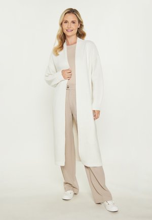 Cardigan lungo bianco in maglia testurizzata con apertura frontale e spalle abbassate. Abbinato a un top beige e pantaloni chiari, su uno sfondo neutro.