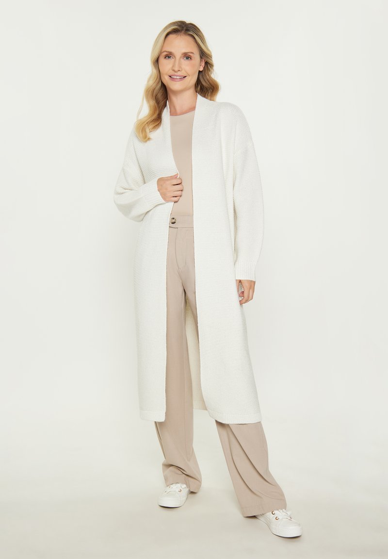 Cardigan lung alb din tricot texturat, cu deschidere în față și umeri lăsați. Asortat cu un top bej și pantaloni deschiși, pe un fundal neutru.