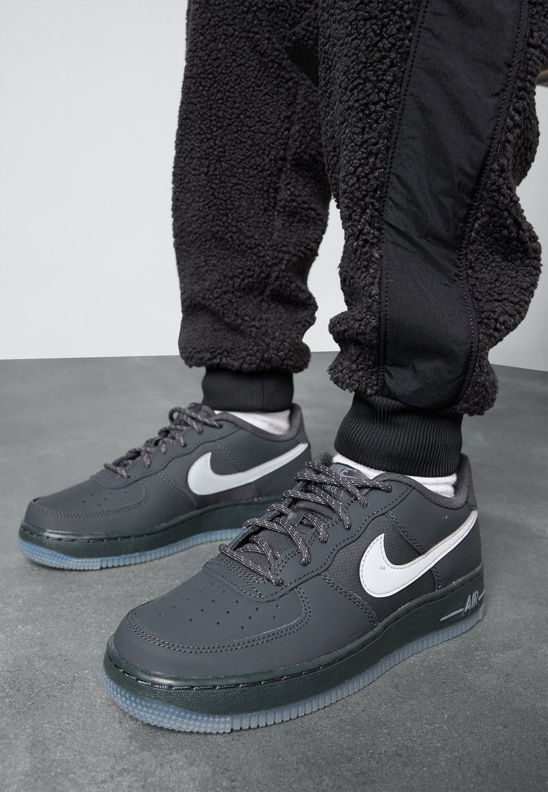 Baskets Nike Air Force 1 noires avec le swoosh blanc, portées avec un pantalon en polaire noir et des chaussettes blanches sur un sol gris.