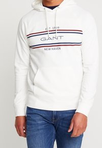 Felpa bianca con cappuccio, con accenti a strisce blu navy e bordeaux, dotata di tasca a marsupio frontale e grafica "GANT NEW HAVEN".