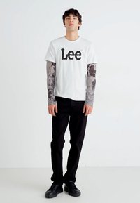 Hvit t-skjorte med svart "Lee" logo, lagd med grå mønstrede langermede skjorter. Svart bukser og svarte joggesko fullfører antrekket.